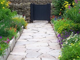Landscaping stones A sidewalk made of landscaping stones surrounded by bright colorful flowers and greenery leading up to a black gate.