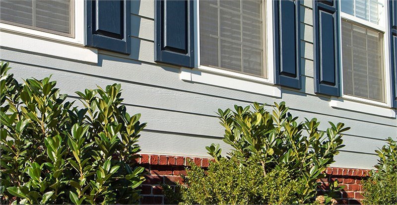 The side of a home with gray lap siding and red brick on the lower half of the siding. There are green hedges in front of the home.