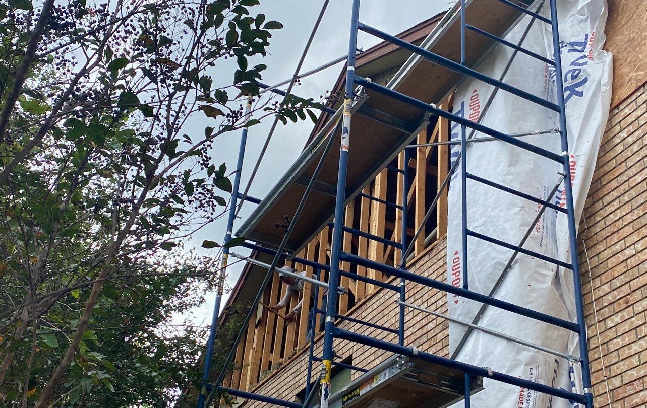 The side of a house with red brick and the top of the house frame exposed. There is a tarp hanging off the side of the house and a scaffold build along the side. Tree branches are covering the left side of the frame.