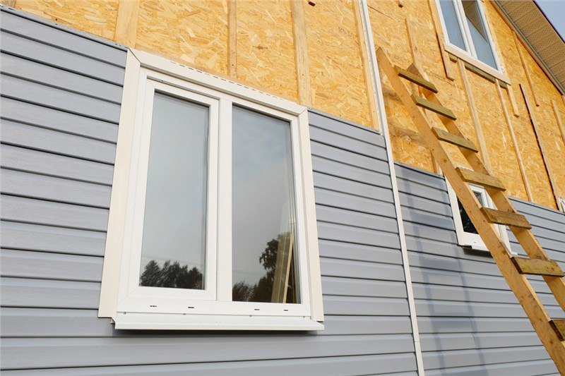 A home having the siding installed with half of the substrate exposed and half of the home with gray siding installed. There is a white window on the left side of the home and a wooden ladder against the home.