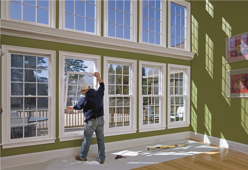A man in a living room with a wall full of windows holding the bottom sash of one window open.