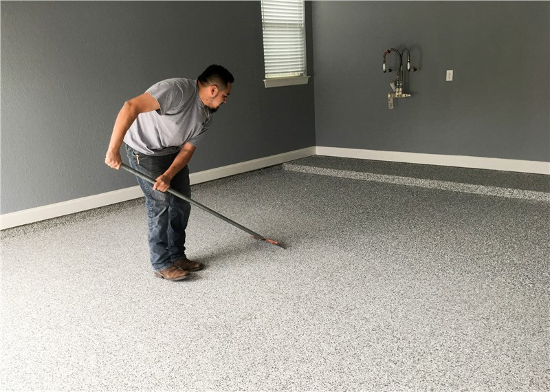 A man in dark pants and a grey tshirt brushing a concrete surface with a broom inside of an empty garage.