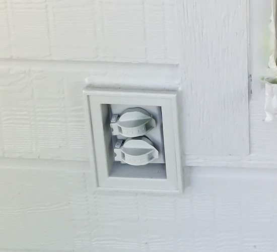 A close up of an outdoor electrical outlet on the side of a home with white siding. 