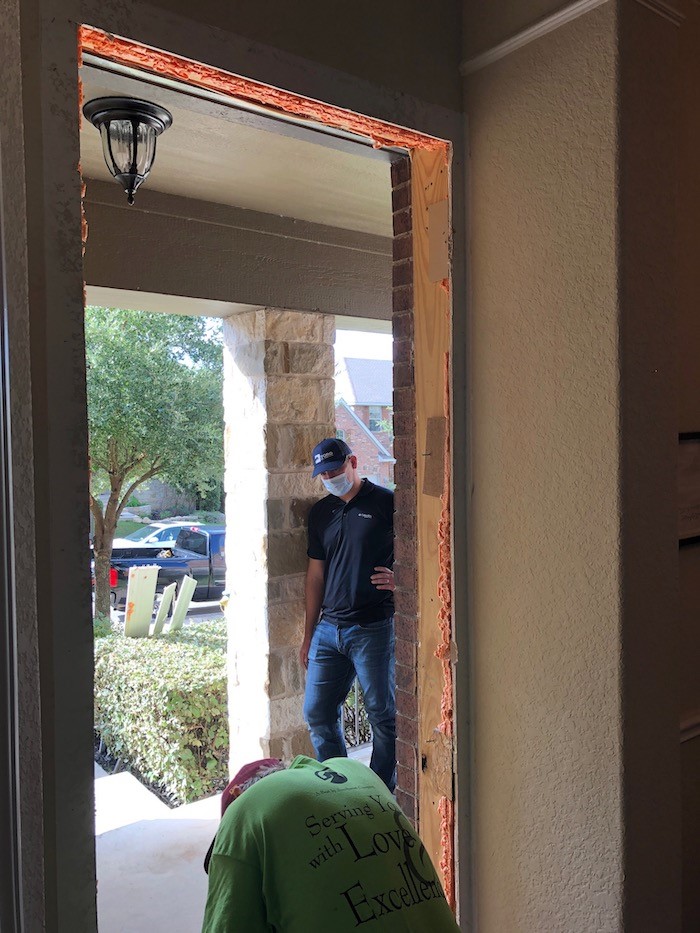 A man in a bright green shirt bent down at the base of a door frame looking outside of the house where a man wearing black is standing looking towards him. The door is removed from the frame and empty.