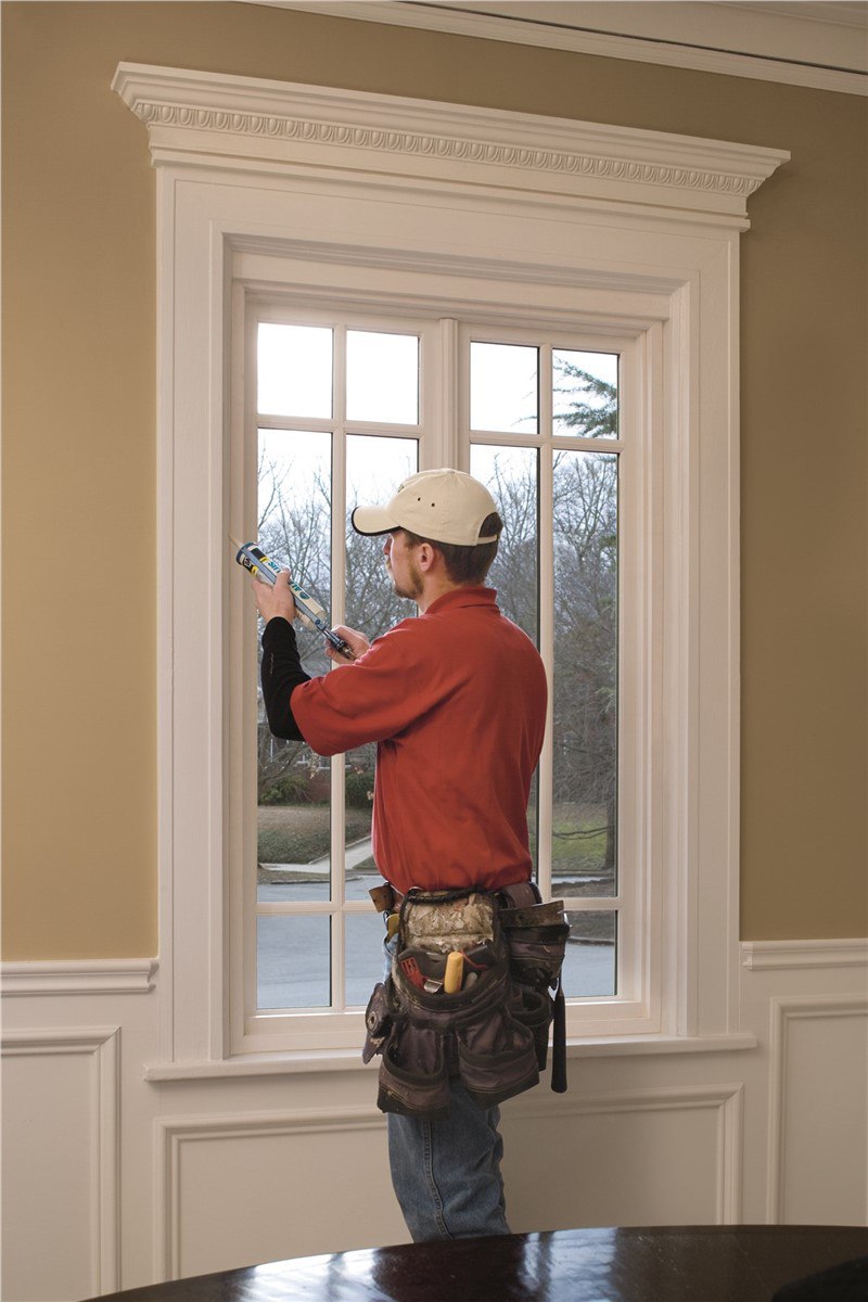 A man caulking the inside of a white window. The window has gridding details and is tall.